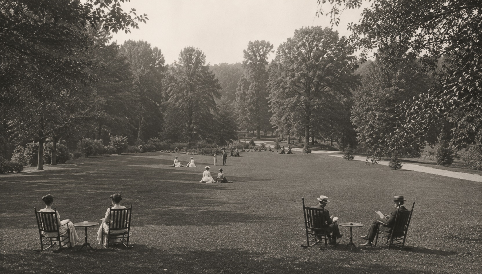 Guests relax on a landscaped lawn beneath mature trees in the early 1900s.
